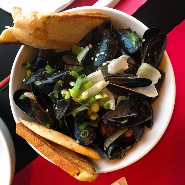 A white bowl of mussels with sliced onions, green onions, and toasted bread on a red table.